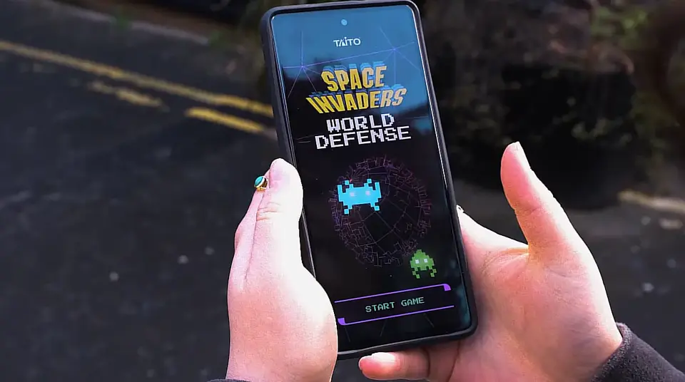 A person holding a smartphone displaying the game 'Space Invaders World Defense' by Taito. The screen shows pixelated alien characters with the words 'START GAME' at the bottom. The background reveals a blurred street scene.