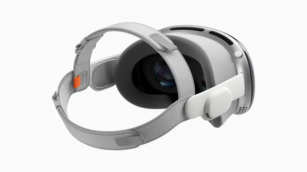 Apple Vision Pro headset featuring a sleek, modern design with a predominantly white color scheme. The headset has an adjustable strap for a secure fit and an oversized, prominent lens at the front for immersive viewing. The side of the device includes an orange button, likely for power or function control, and there's a touch-sensitive interface on the side arm. The build appears sturdy with a combination of smooth and textured surfaces.