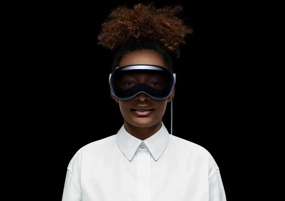 A woman wearing the Apple Vision Pro headset, featuring a sleek design with a wide headband, stands against a black background. She is dressed in a crisp white shirt, suggesting a professional or cutting-edge atmosphere.